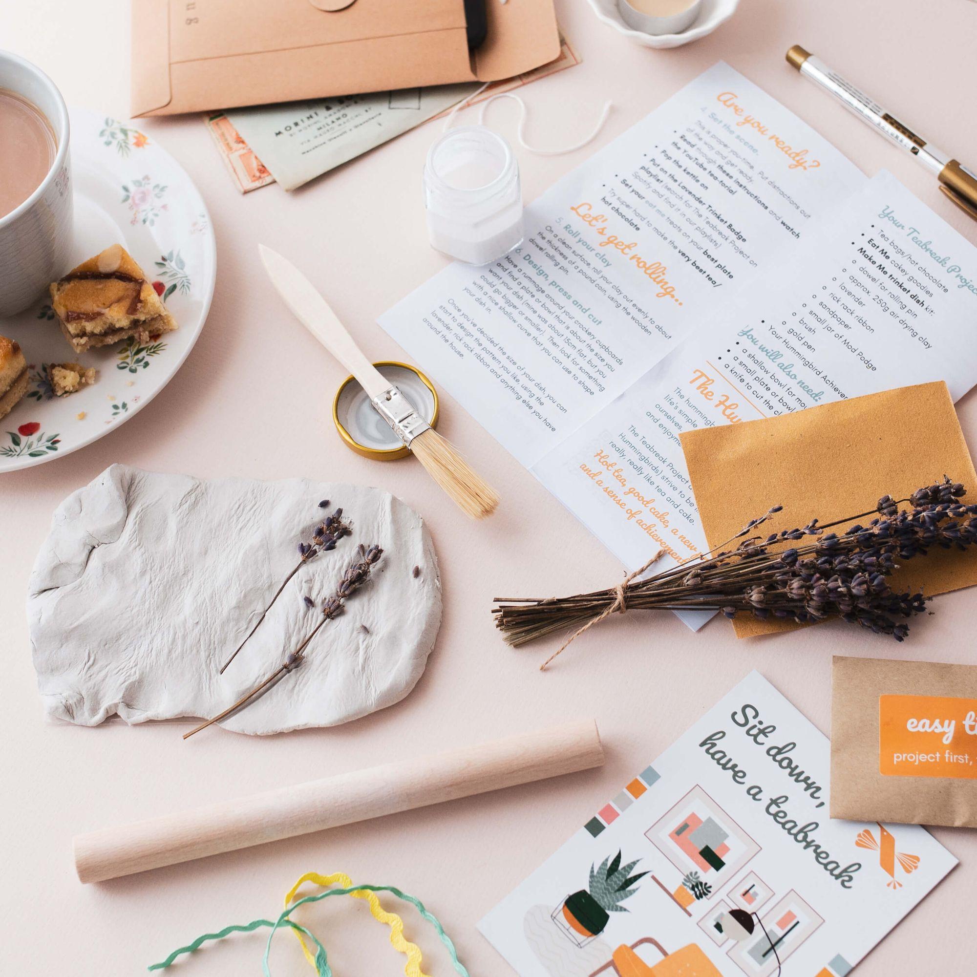 A clay trinket dish being made on a kitchen table, with lavender, a rolling pin, project instructions, a mug of tea and a slice of cake