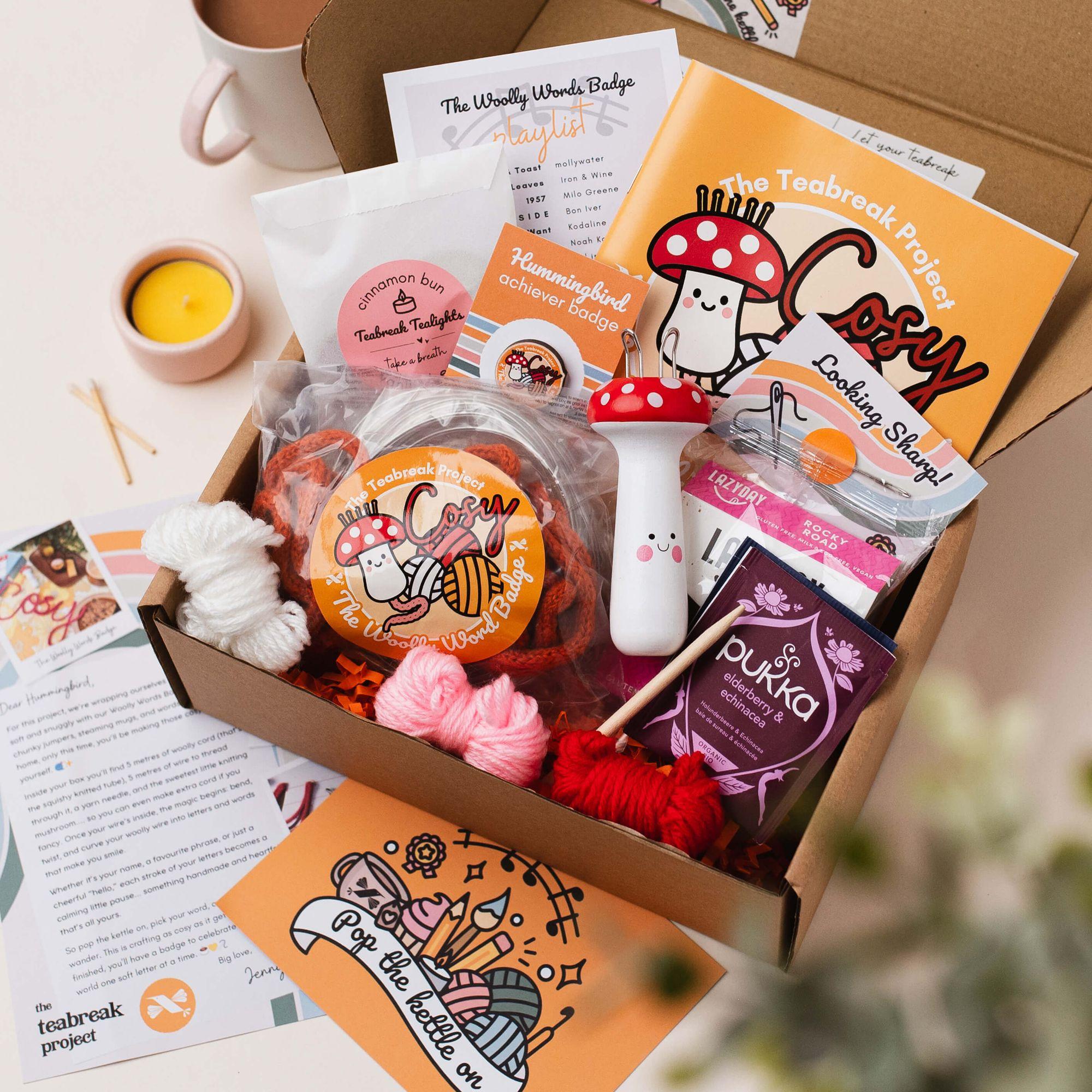 An opened Teabreak box on a kitchen table holding a knitting project, mushroom tool, tea, a gluten-free brownie, a playlist card and a Hummingbird badge